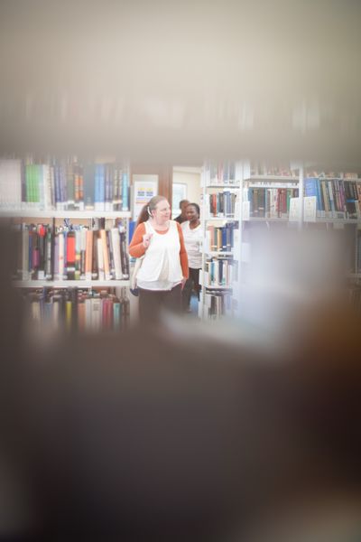 Students walking between bookshelves in a library, viewed through rows of books.
