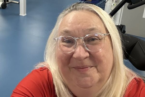 Angela, a blonde woman in her late 50s, smiles at the camera. She is in the gym at the hospital where she is being rehabilitated following a spinal injury.