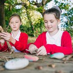 two primary aged girls sat outside on a table and bench making crosses from sticks