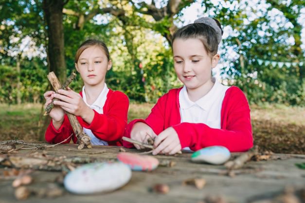 two primary aged girls sat outside on a table and bench making crosses from sticks