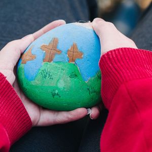 A child's hand holding a painted pebble of the three crosses