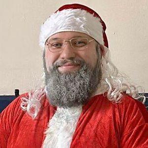 Rev'd Darren Lynch is smiling, dressed in a red santa suit. He has sprayed his beard grey.