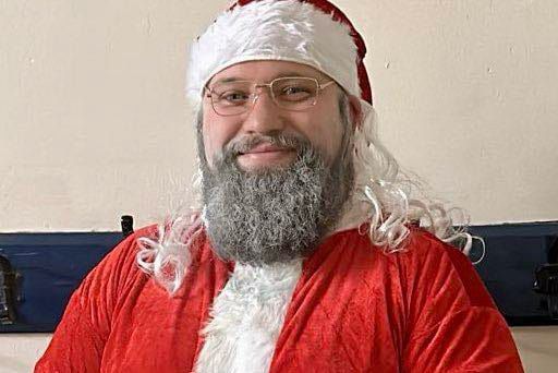 Rev'd Darren Lynch is smiling, dressed in a red santa suit. He has sprayed his beard grey.